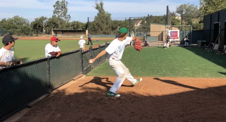 Pitcher winds up to throw to catcher while campers and coaches watch